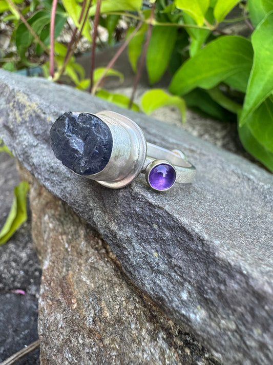 A silver ring with a large dark polished stone and a smaller purple amethyst stone, resting on a gray rock with green foliage in the background.